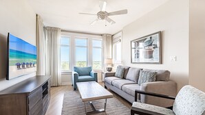 A living room with a flat-screen TV, a ceiling fan, a sofa, a coffee table, and a large window with curtains. - Balcony ~ Picturesque Windows ~ Outdoor Seating (Miramar Beach)