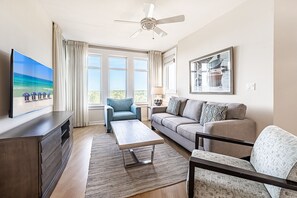 Un salón amplio con una televisión de pantalla plana grande, un ventilador de techo, un sofá, una mesa de centro y un ventanal con vistas al exterior.