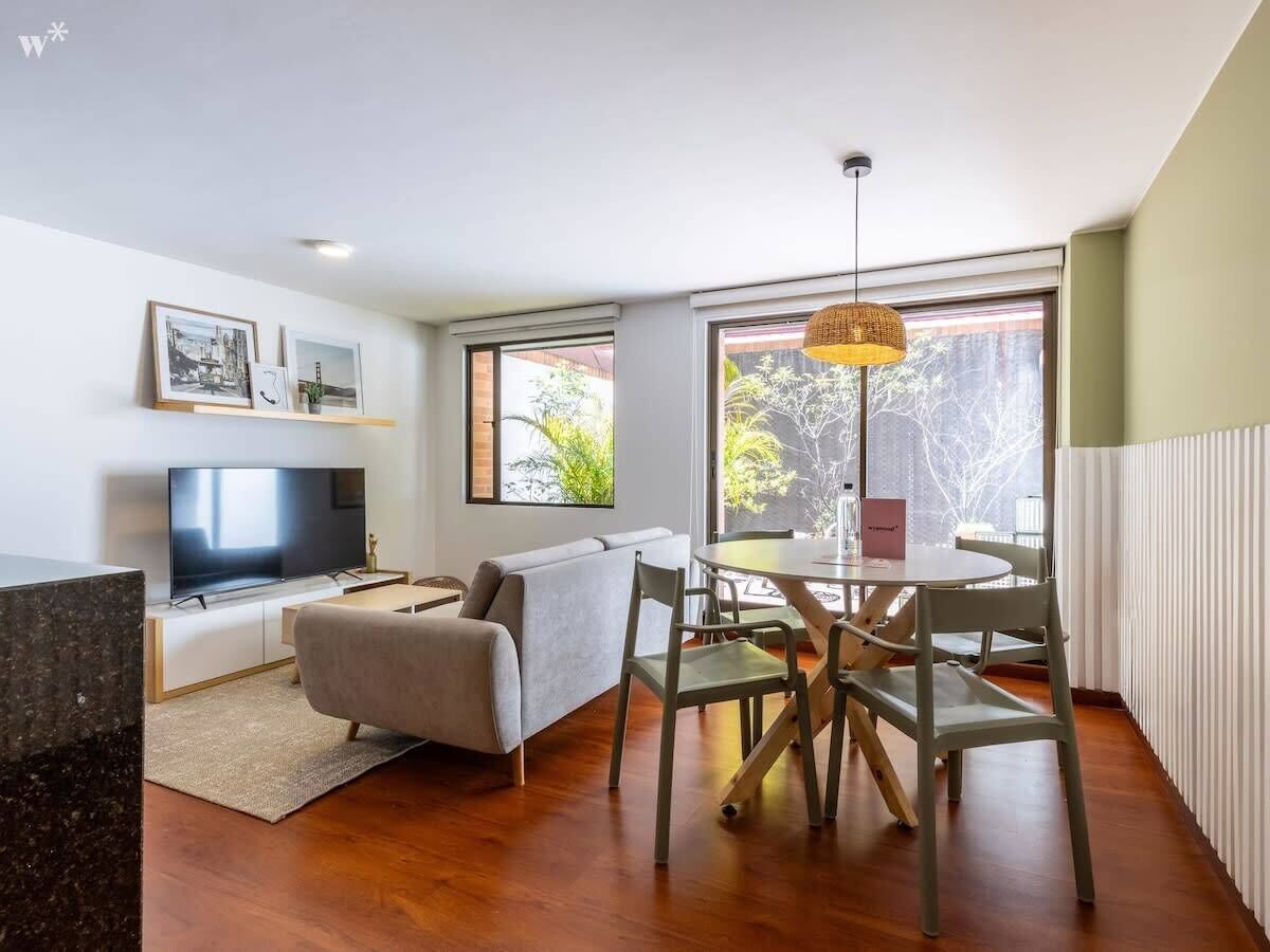 A modern living room with a flat-screen TV, a sofa, a dining table with chairs, and large windows with a view of greenery.