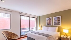 Ein Schlafzimmer mit einem großen Bett, einem Sessel, einem Nachttisch und einem Fenster mit Blick auf ein Gebäude.