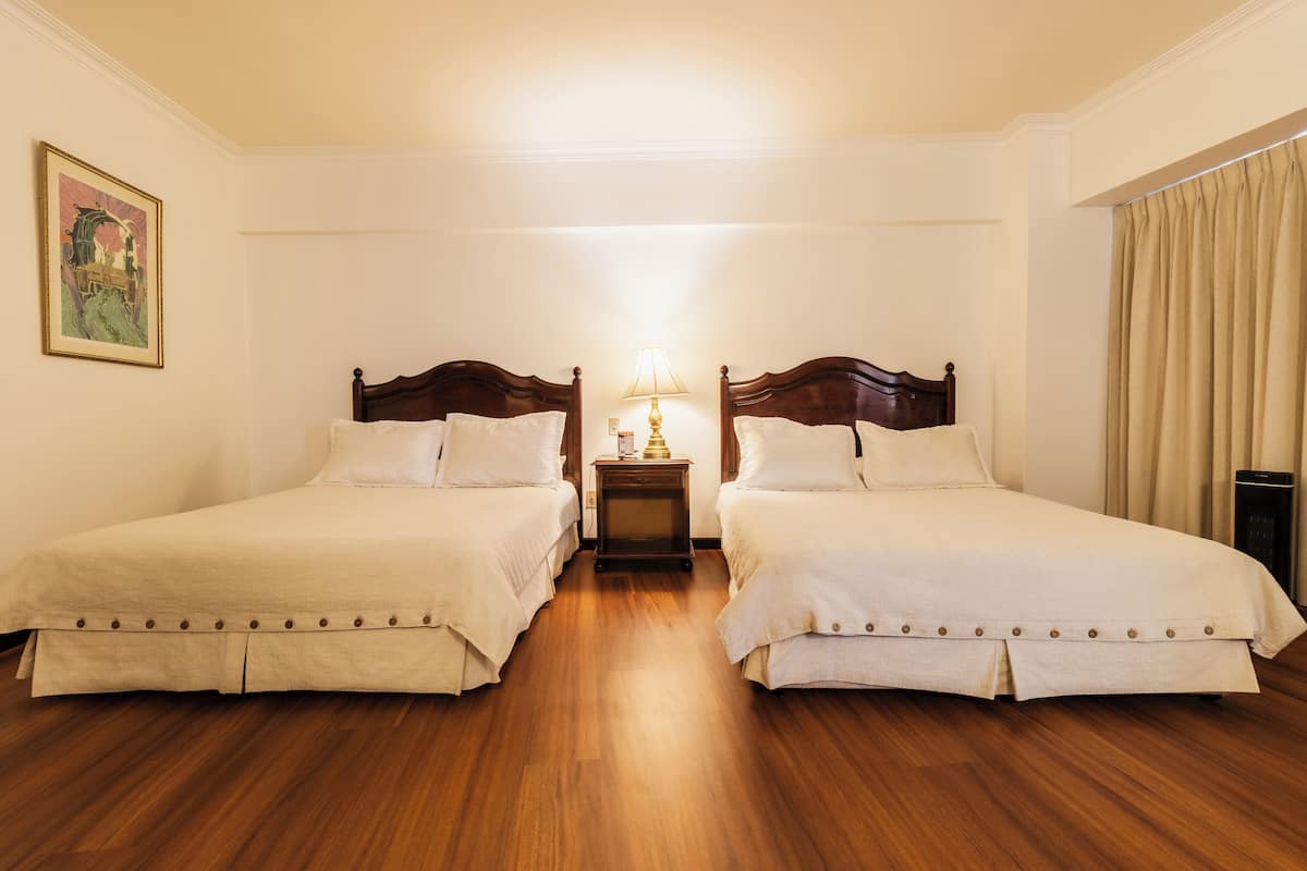 Habitación de hotel con dos camas individuales, una mesita de noche con lámpara, suelo de madera y un cuadro en la pared.