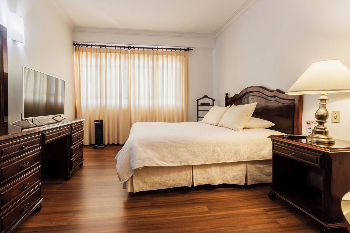 Habitación de hotel ordenada con una cama grande, una mesita de noche con lámpara, un aparador y un televisor sobre un mueble.