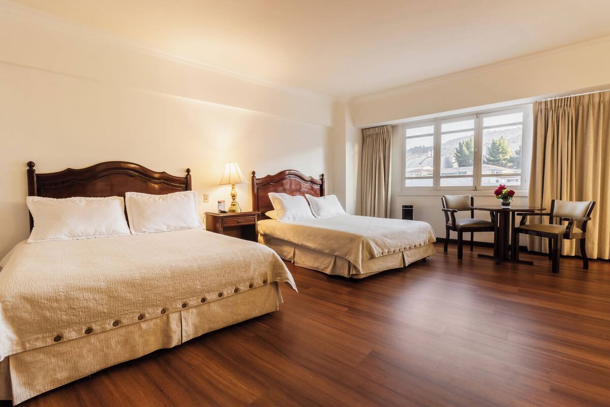 Habitación de hotel con dos camas, piso de madera, una mesa de comedor con sillas y una ventana con cortinas.