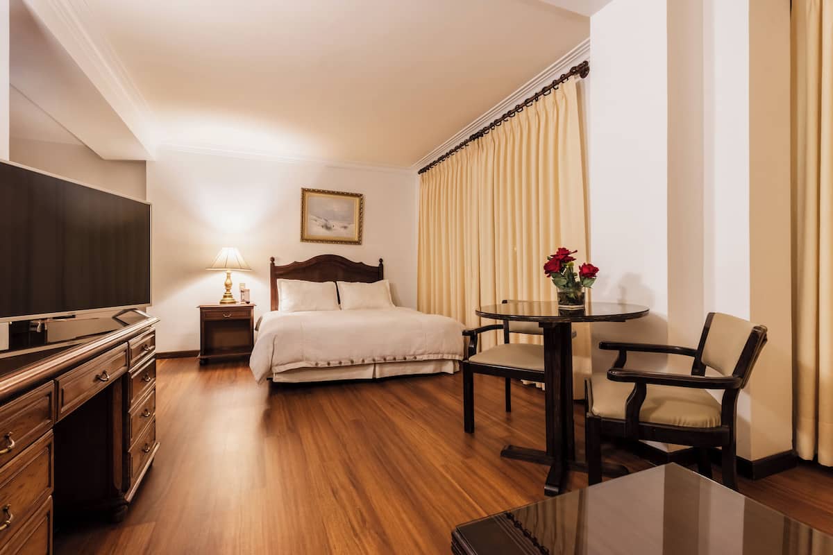 Una habitación de hotel con una cama, un televisor, una mesita de noche con una lámpara, una pequeña mesa con un jarrón de flores y una silla.