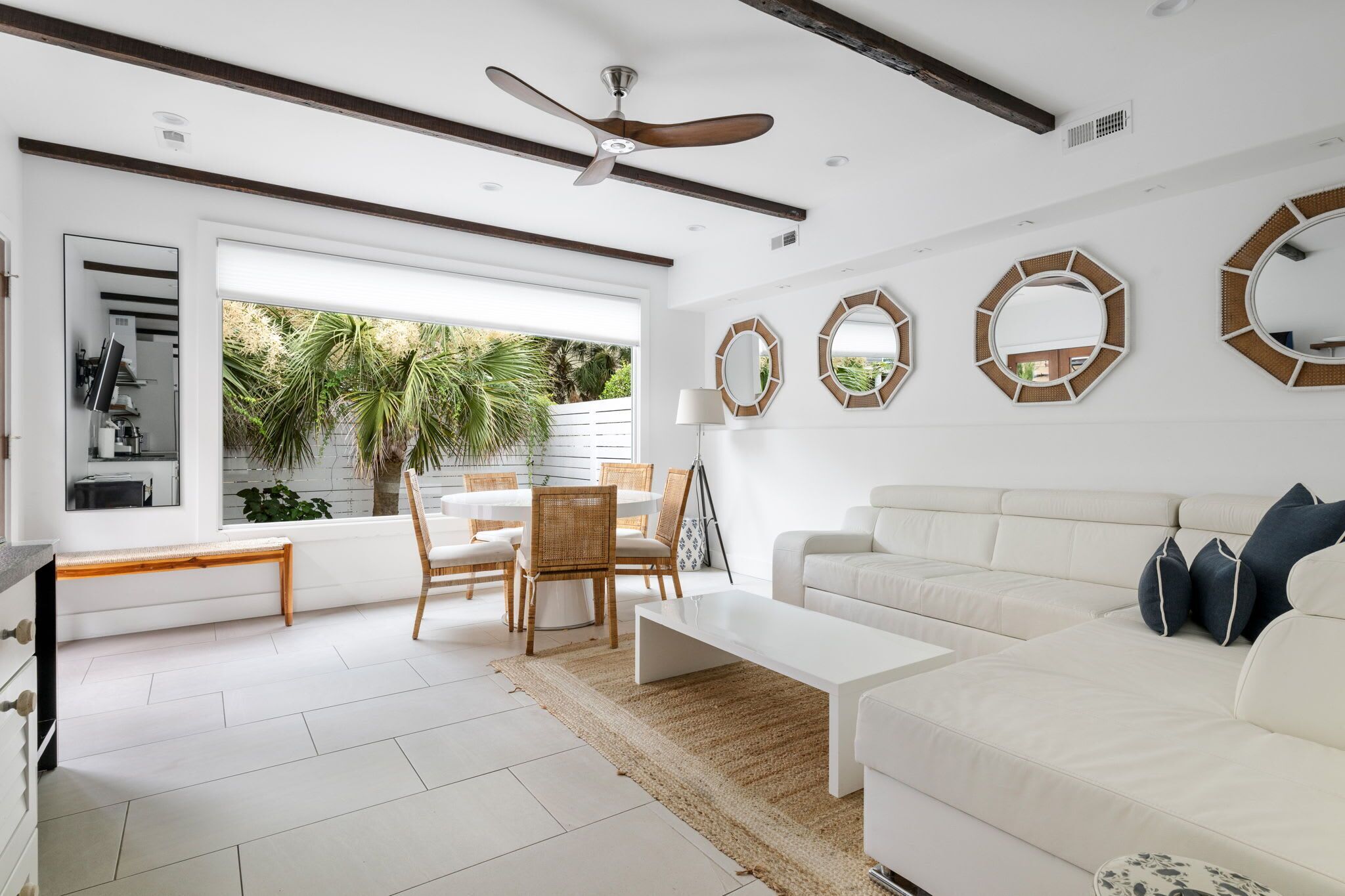A modern living room with a white sofa, a dining table with chairs, a ceiling fan, and large windows with a view of palm trees.