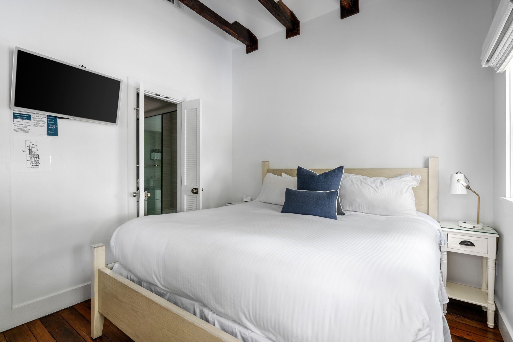 Un dormitorio con cama, mesita de noche, lámpara de escritorio, televisor de pantalla plana y puerta que da a un baño.