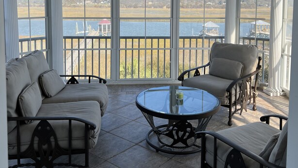 A covered outdoor seating area with a ceiling fan, large windows, and a view of a body of water and distant buildings. - Luxury Waterfront Paradise at Ocean Isle Beach: Enjoy the Beach and Jink's Creek (Ocean Isle Beach)