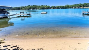 A clear lake with a sandy beach, a dock with a boat, and a row of houses in the background. - Whitewater Lakefront Lodge with 200 Feet Shoreline Docks Sunset Views (Whitewater)