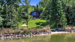 A lakeside lodge with a large deck, surrounded by dense trees and a well-maintained lawn. - Whitewater Lakefront Lodge with 200 Feet Shoreline Docks Sunset Views (Whitewater)