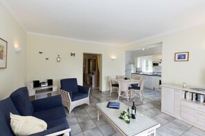 Un salon comprenant un canapé, un fauteuil, une table à manger et des chaises. On aperçoit une cuisine au fond.