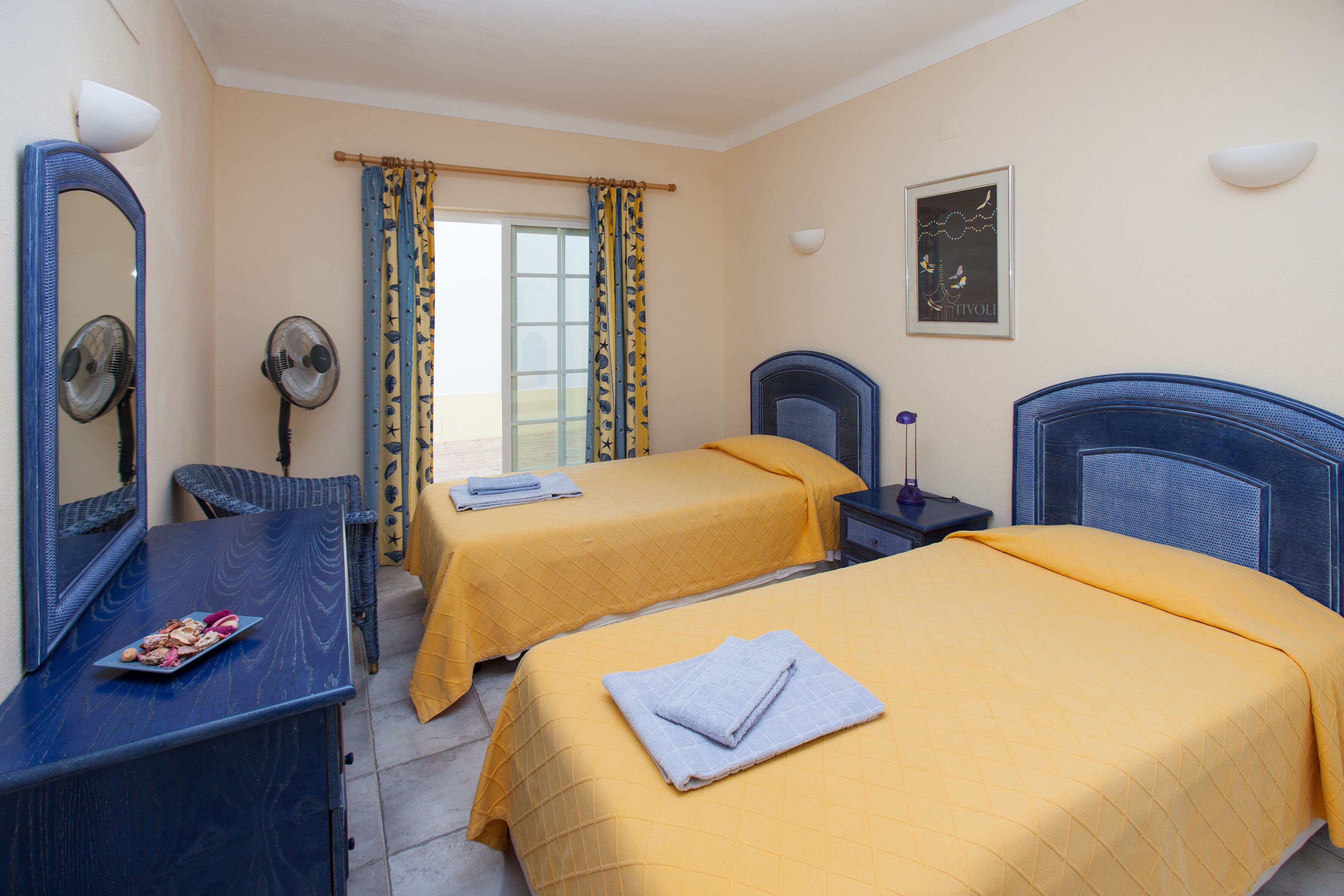 Une chambre d’hôtel avec deux lits, un bureau, une chaise, un miroir, un ventilateur et une fenêtre avec des rideaux.