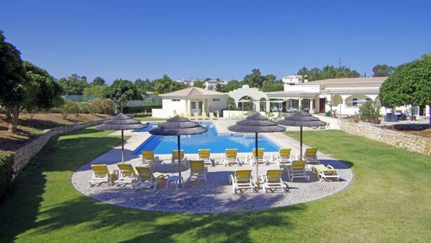 Zona de piscina con sillones de descanso y sombrillas, rodeada de un césped cuidado y un edificio con techo de tejas.