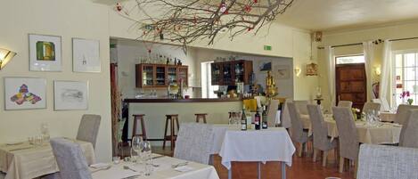 Un restaurante con varias mesas y sillas, una zona de bar con estantes llenos de botellas, y una rama decorativa colgando del techo.