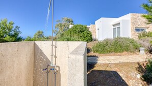 A modern outdoor shower area with a stone wall, a clear sky, and a building with a stone facade in the background. - Superb villa with pool and 180° sea view (Torre Pali)