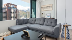 A modern living room with a grey sectional sofa, a black coffee table, and a small round table with a patterned table runner. A framed picture hangs on the wall, and a large window offers a view of city buildings and mountains. - 22nd Floor Apartment  With Mountain Views (Cape Town)