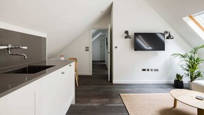 Eine moderne Küche mit dunkler Arbeitsplatte, weißen Schränken und einer eingebauten Spüle. Ein Fernseher ist an der Wand montiert und rechts steht eine Pflanze.