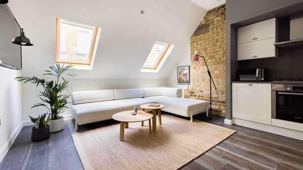 Ein modernes Wohnzimmer mit einer Couch, zwei kleinen Tischen, einem großen Fenster, einer Ziegelwand und einer Küchenzeile.