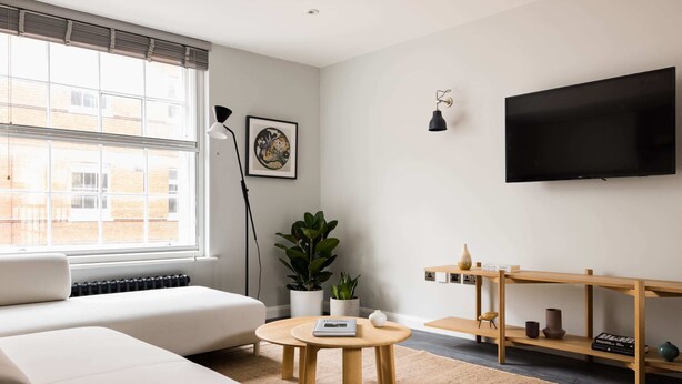 Ein modernes Wohnzimmer mit einer Couch, einem runden Holztisch, einem Blumentopf, einem Bücherregal und einem wandmontierten Flachbildfernseher.
