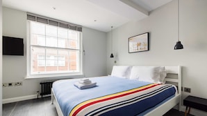 Ein modernes Schlafzimmer mit großem Fenster, einem Bett mit gestreifter Decke, einem Nachttisch und einem an der Wand montierten Fernseher.