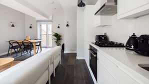A modern kitchen with white cabinetry, a gas stove, and a dining area with a white sofa and wooden table. - Marvellous Leicester Sq 2BR - Netflix & Nespresso (London)