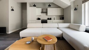A modern living room with a sectional sofa, a wooden coffee table, and a kitchen area with white cabinets and black appliances. - Marvellous Leicester Sq 2BR - Netflix & Nespresso (London)