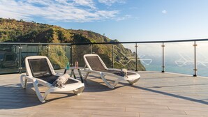 A sunlit outdoor lounge area with two lounge chairs, a glass barrier, and a view of a coastal landscape. - Modern villa, shared infinity pool | SunsetCliff 4 (Faja Da Ovelha)
