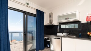 A modern kitchen with a dishwasher, oven, and microwave. It has a large window with blue curtains and a view of the sea. - Modern villa, shared infinity pool | SunsetCliff 7 (Faja Da Ovelha)