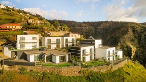A modern residential area with contemporary houses, a stone retaining wall, and a hillside backdrop. - Modern villa, shared infinity pool | SunsetCliff 3 (Faja Da Ovelha)