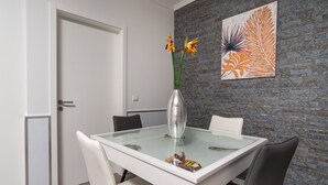 A dining area with a glass-top table, white chairs, and a vase with flowers. - Modern villa, shared infinity pool | SunsetCliff 3 (Faja Da Ovelha)