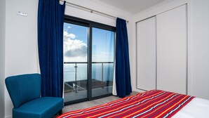 A hotel room with a large bed, a blue chair, a balcony with a glass door, and a white wardrobe. - Modern villa, shared infinity pool | SunsetCliff 3 (Faja Da Ovelha)