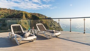 Une terrasse dotée de deux chaises longues, d’une balustrade en verre et offrant une vue sur un paysage côtier.