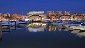 Un puerto deportivo con barcos amarrados, un edificio moderno de techo plano y un paisaje urbano con diversos edificios al fondo.