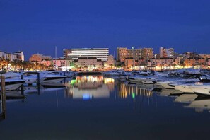 A marina with boats docked, a modern building with a flat roof, and a cityscape with various buildings in the background.