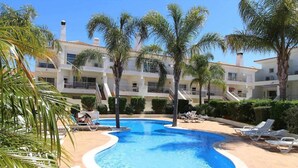 Una piscina con sillas de descanso, palmeras y un edificio de varios pisos con balcones.