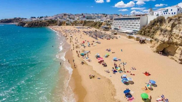 Una playa con una hilera de edificios blancos, cielo despejado y mar tranquilo.
