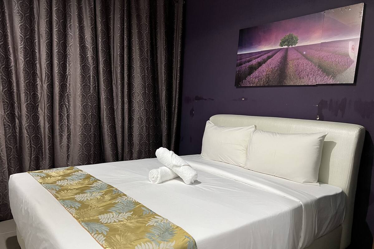 A neatly made bed with a floral bedspread, a white headboard, and a painting of a landscape on the wall.