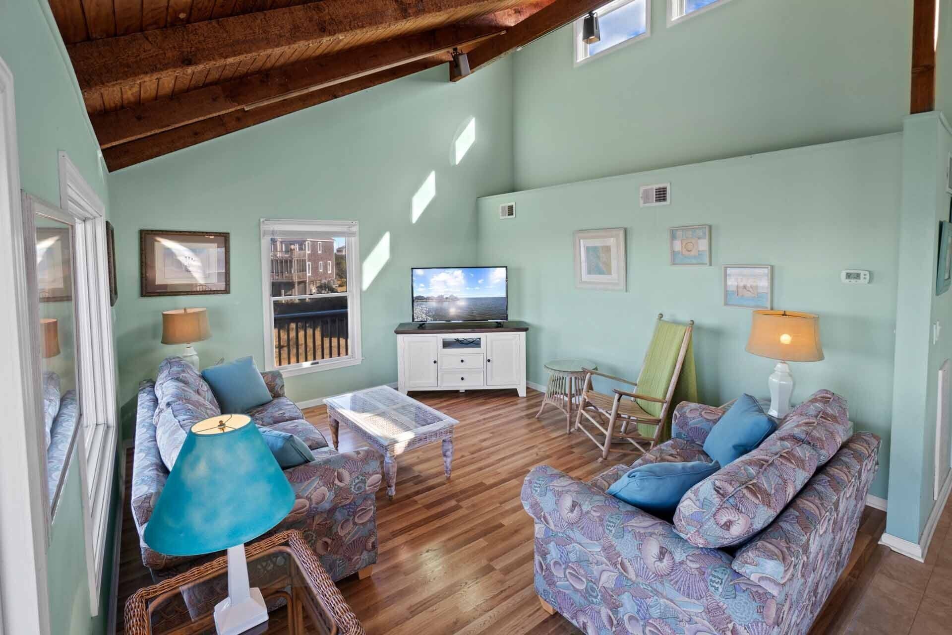 A spacious living room with a high ceiling, a television, a dining area with a table and chairs, and a view through a window.