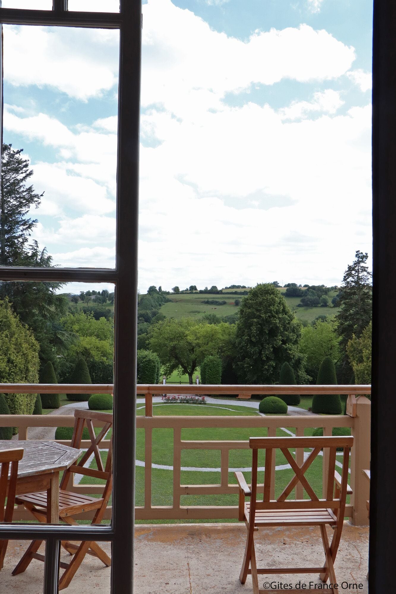 A balcony with a wooden table and chairs, overlooking a green landscape with trees and a clear sky.