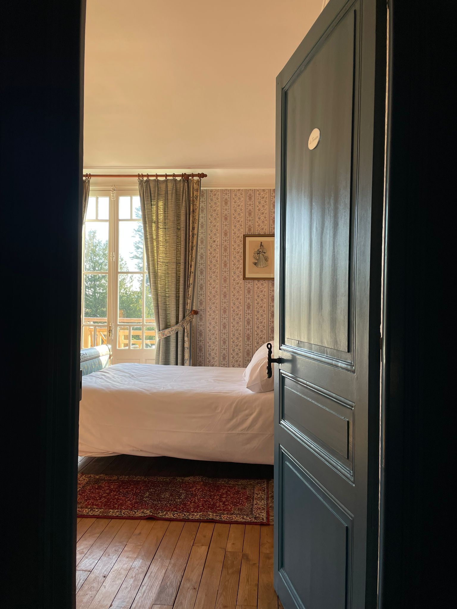 A bedroom with a bed, a window with curtains, a door, and a patterned wallpaper.