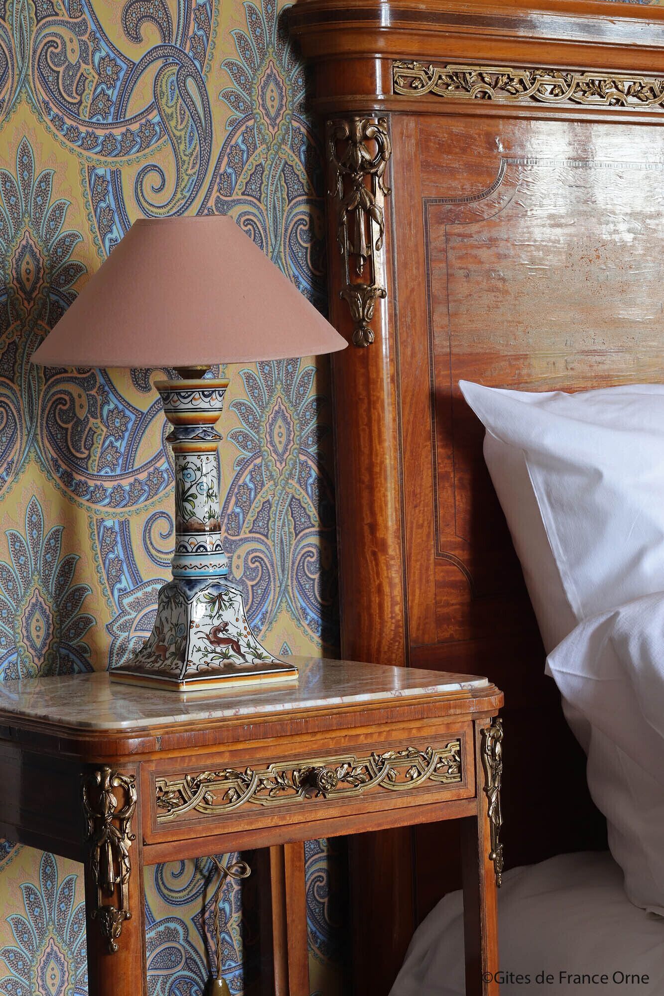 A wooden bedside table with a decorative lamp and a book, against a wall with a paisley-patterned wallpaper.