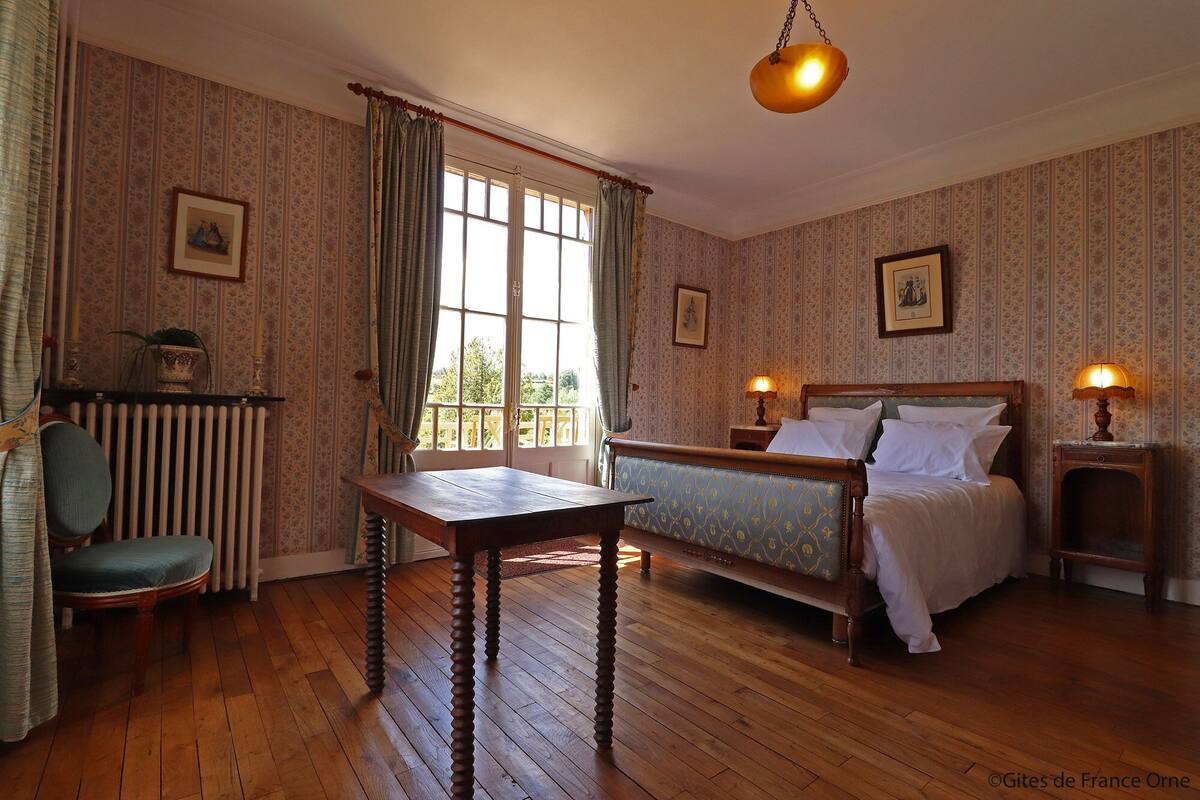 Een kamer met houten vloer, een bed met hoofdeinde, een tafel, een stoel, een radiator en een raam met gordijnen.
