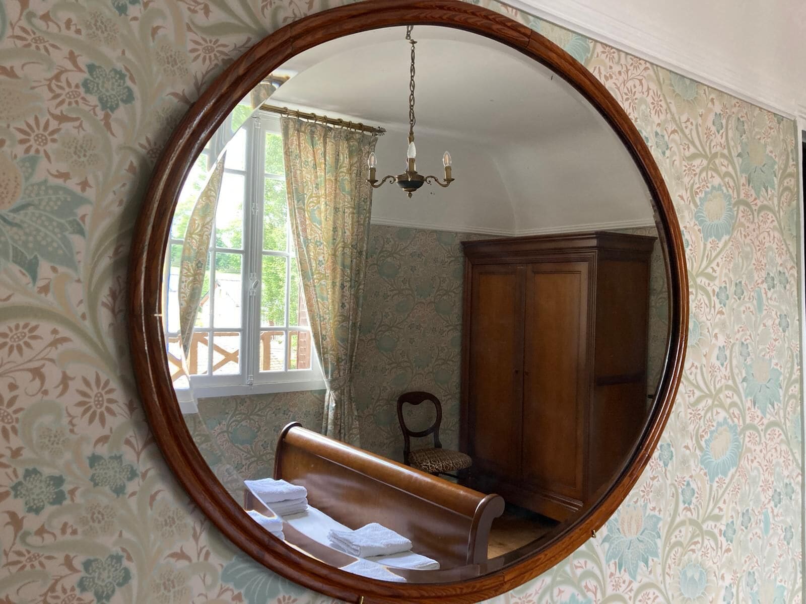 A room with a large round mirror, a wooden bench with towels, a wooden cabinet, and a window with curtains.