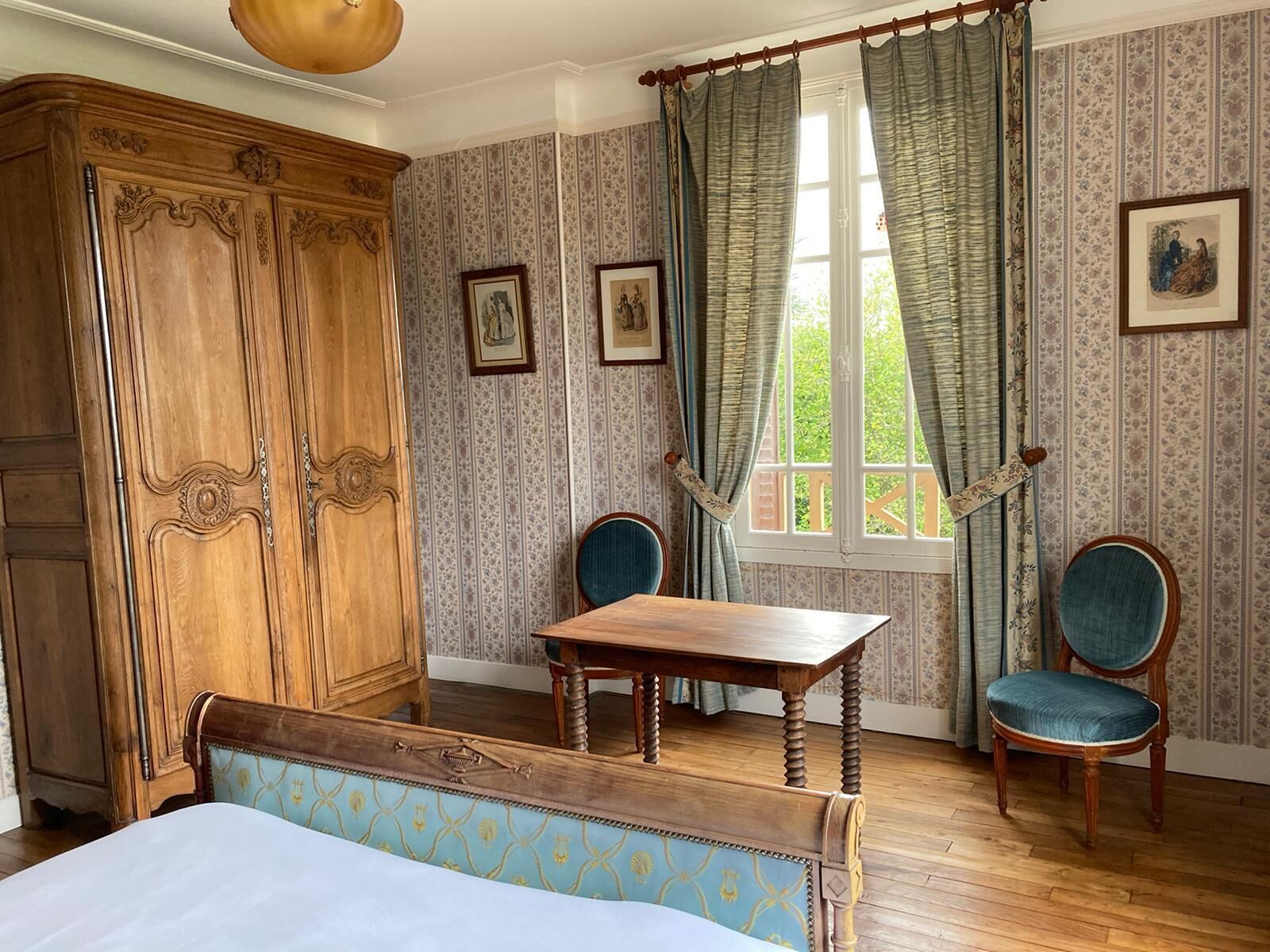 A room with a wooden wardrobe, a bed with a patterned headboard, a wooden table, two chairs, and a window with curtains.