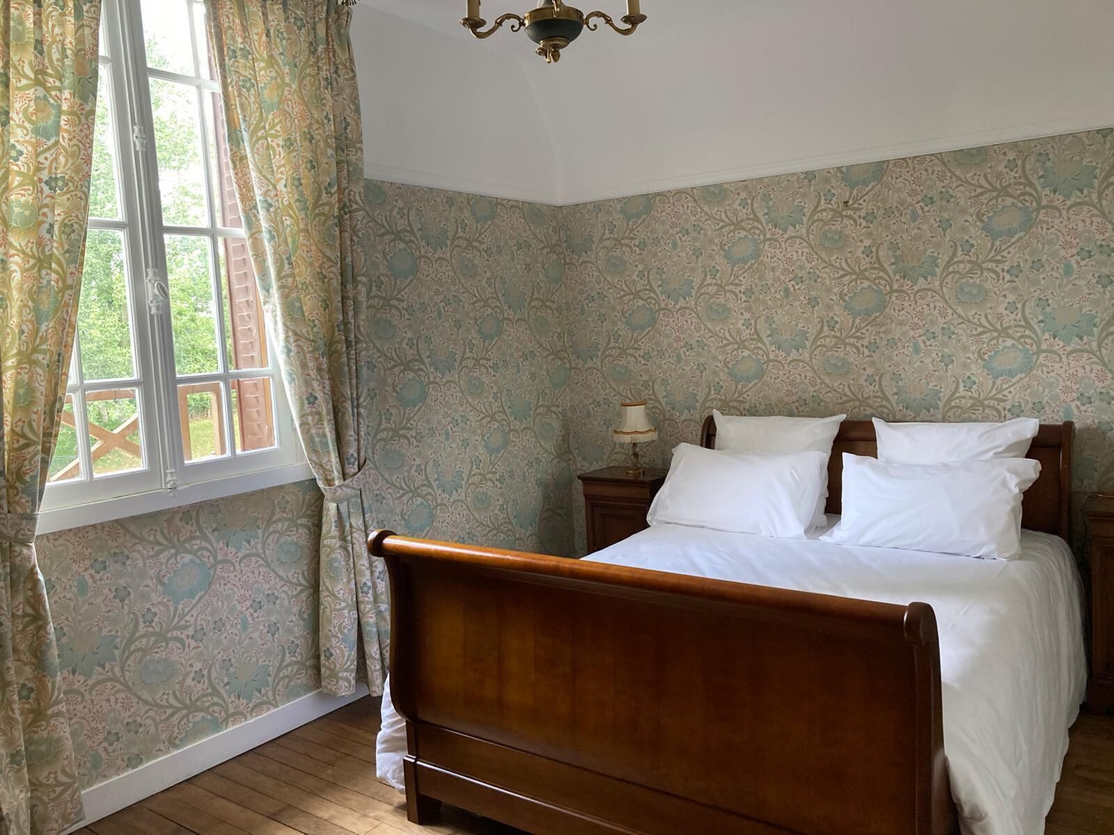 Un dormitorio con cama de madera, papel tapiz estampado, una ventana con cortinas y un candelabro.