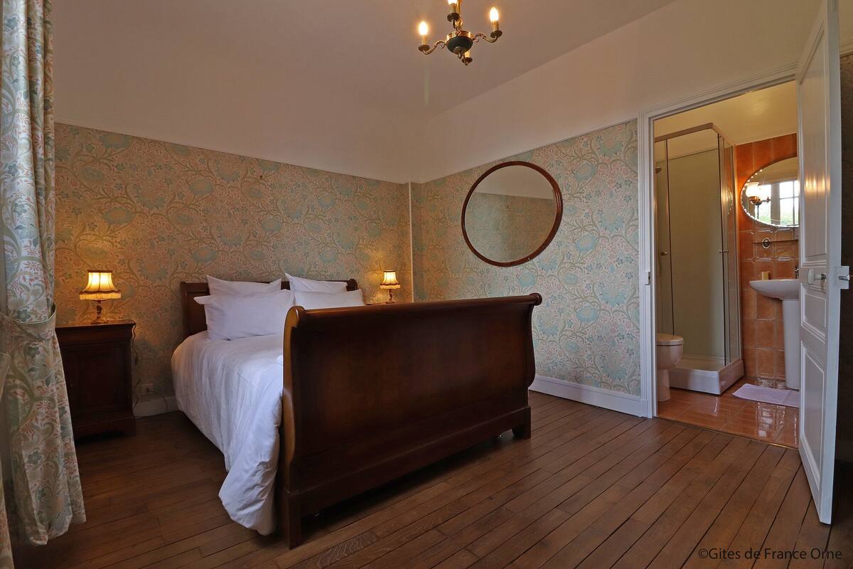 Een slaapkamer met een houten bed, twee bedlampen, een ronde spiegel en een badkamer zichtbaar door een geopende deur.