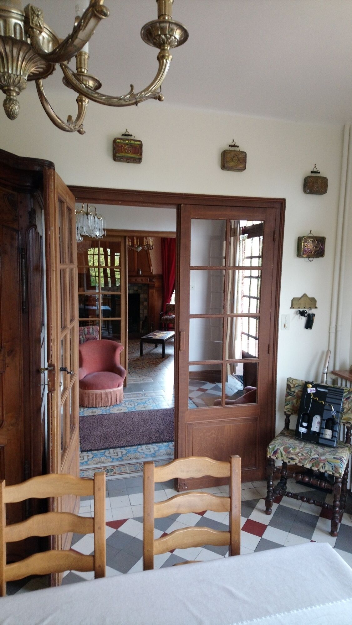 Una habitación con piso a cuadros, una silla de madera, un sillón rojo, una araña y una puerta que da a otra habitación.