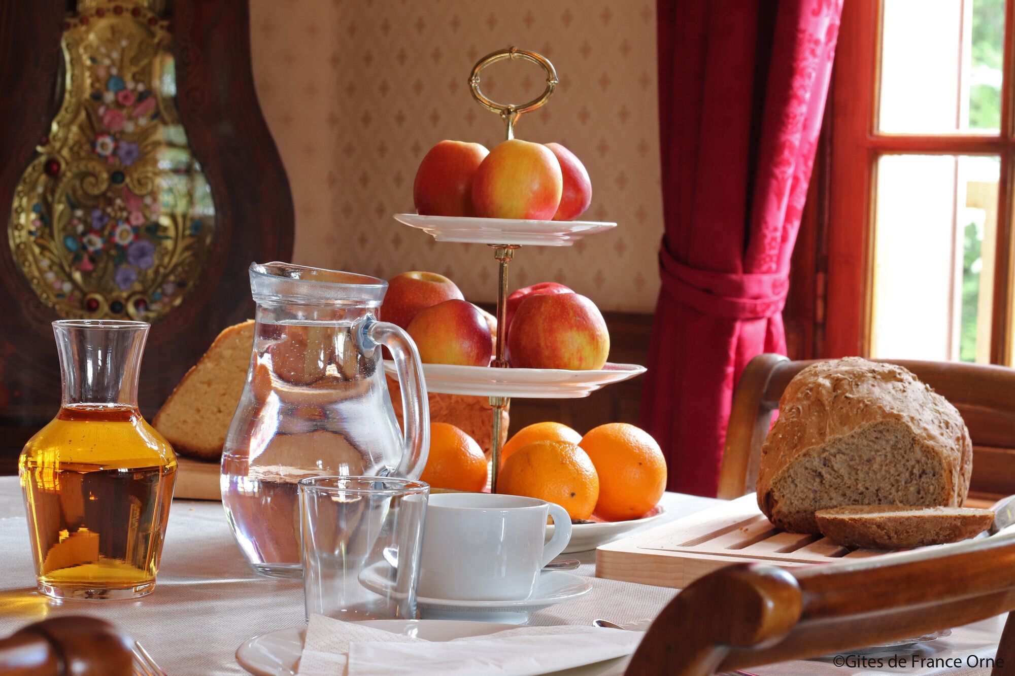 Une table mise pour le petit-déjeuner avec un pichet, des tasses, des oranges, du pain et une assiette décorative ornée de fruits.