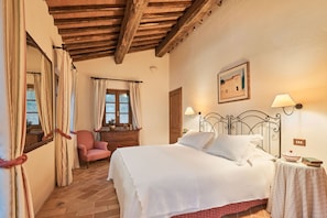 A bedroom with a wooden ceiling, a bed with white bedding, a chair, a bedside table with a lamp, and a window with curtains.