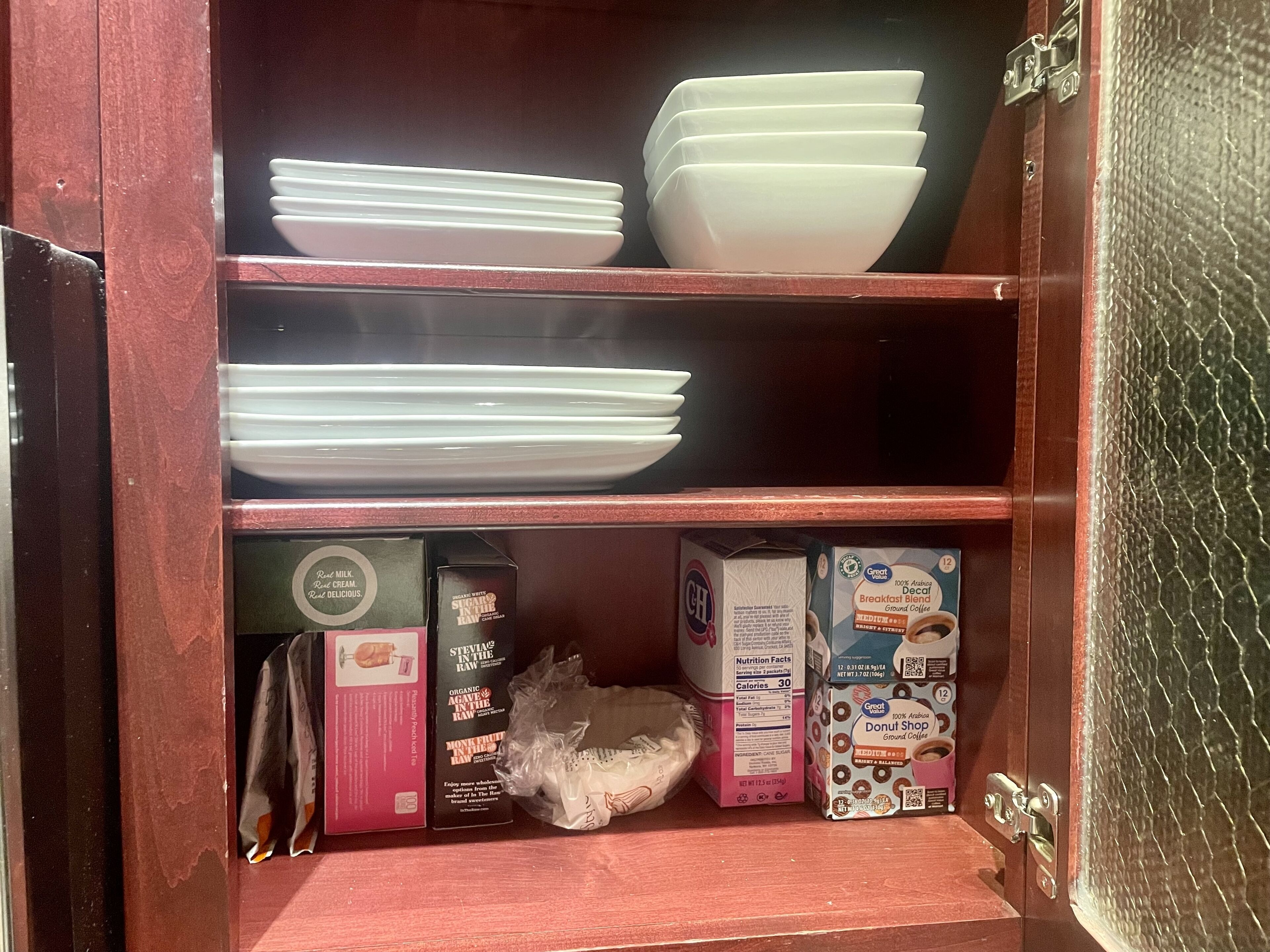A wooden cabinet with shelves containing white plates, a box of Red Stripe beer, a box of Monk Fruit in the Raw, a box of Great Value Decaf Breakfast Blend, and a box of Great Value Donut Shop Ground Coffee.