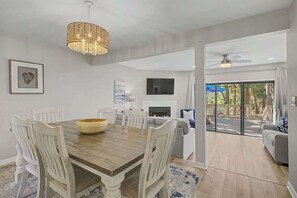 A dining area with a wooden table, white chairs, a woven bowl, a hanging light fixture, and a TV mounted on the wall.